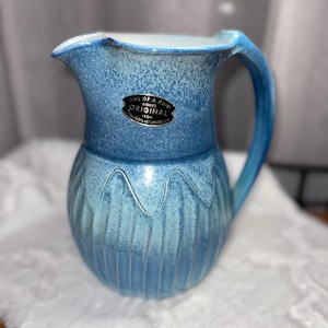 Könnte beinhalten: Ein blauer Keramikkrug mit Henkel und Ausguss. Der Krug hat ein strukturiertes, geripptes Design auf der unteren Hälfte und eine glatte obere Hälfte. Ein Etikett lautet "ONE OF A KIND SIGNED ORIGINAL".