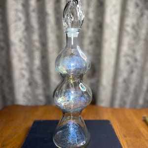 May include: A clear glass bottle with a decorative stopper. The bottle has a unique shape with three bulbous sections and iridescent coloring. The stopper is a clear, teardrop-shaped design. The bottle is sitting on a dark blue book.