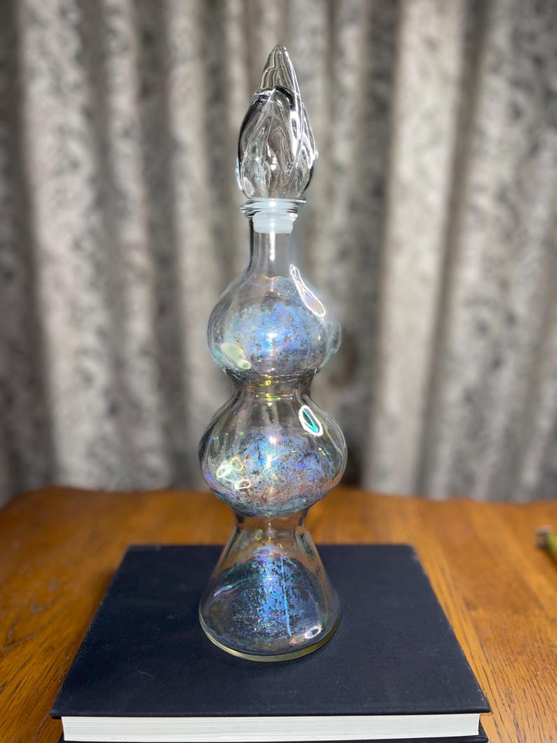 May include: A clear glass bottle with a decorative stopper. The bottle has a unique shape with three bulbous sections and iridescent coloring. The stopper is a clear, teardrop-shaped design. The bottle is sitting on a dark blue book.