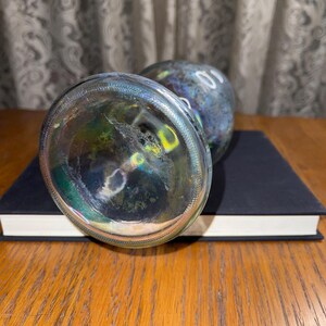 May include: Two glass bottle stoppers with iridescent colouring, resting on a closed book. The stoppers have a rounded shape and a clear, textured base. The book is dark blue with white-edged pages.