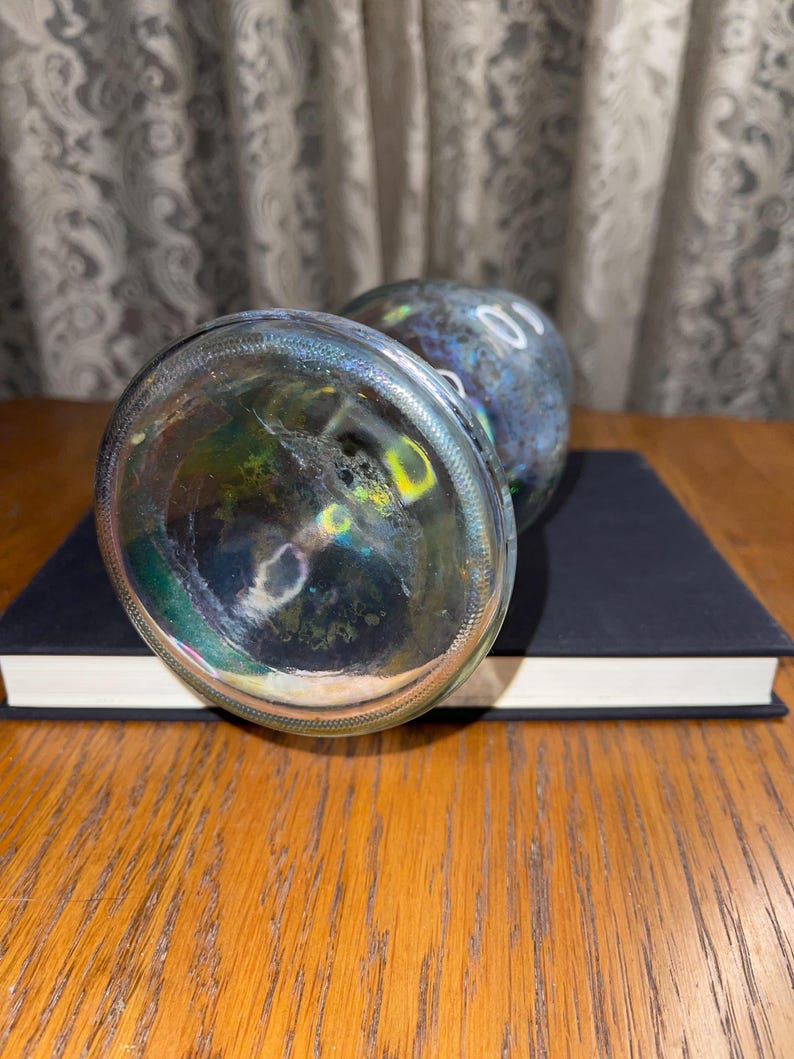 May include: Two glass bottle stoppers with iridescent colouring, resting on a closed book. The stoppers have a rounded shape and a clear, textured base. The book is dark blue with white-edged pages.