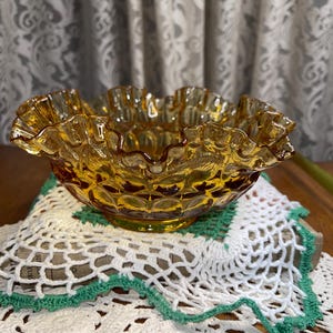 May include: An amber-coloured glass bowl with a ruffled rim. The bowl has a textured, patterned surface and sits on a white and green crocheted doily. A decorative piece.
