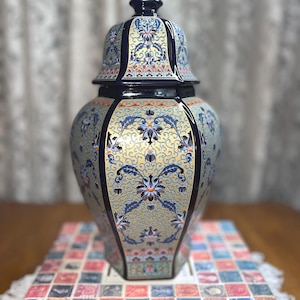 Op de afbeelding: Een decoratieve keramische vaas met een deksel. De vaas is marineblauw met gouden en bloemmotieven. De vaas staat op een boek met een kleurrijk postzegelontwerp.