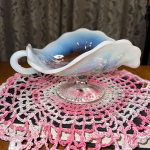May include: An opalescent glass serving dish with a handle, on a pink and white crocheted doily. The dish has a ruffled edge and sits on a clear, pedestal base. The doily is placed on a dark surface.