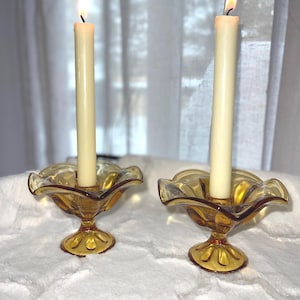 May include: Two lit candles in amber glass candle holders. The candle holders have a scalloped rim and a pedestal base. The candles are ivory-coloured and are lit with a yellow flame. The background is a white surface.