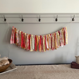 May include: A colorful fabric garland with red, yellow, and white patterned ribbons hangs against a gray wall. The garland is suspended by a thin white string. The image also shows a wooden bowl with yarn, a candle holder, and a small pitcher.