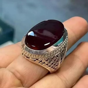 Puede incluir: Un anillo de plata con una piedra preciosa ovalada de color rojo oscuro. El anillo tiene una banda detallada y texturizada y un engaste decorativo alrededor de la piedra. La piedra está pulida y refleja la luz.