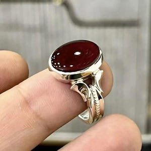 Peut inclure: Bague en argent ornée d'une grosse pierre rouge foncé de forme ovale. La pierre est sertie dans un anneau en argent poli avec des détails ornés sur les côtés. La bague est présentée sur un doigt.
