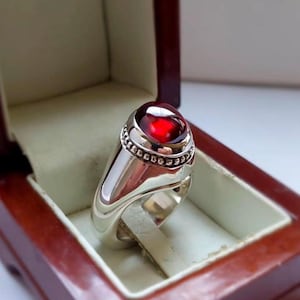 Könnte beinhalten: Ein polierter Silberring mit einem glatten, tiefroten Cabochon-Edelstein, der in einer Fassung mit Perlenrand sitzt. Der Ring hat eine breite, massive Ringschiene, die nach unten hin schmaler wird und eine reflektierende Metalloberfläche aufweist.