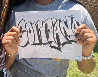 May include: A white sheet of paper with the name "GORIANA" written in black graffiti-style lettering. The letters are outlined in black and filled with white. The paper is held by a person wearing a gray t-shirt.