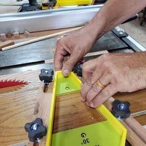 Könnte beinhalten: Eine Person verwendet eine gelb-schwarze Tischsägevorrichtung mit einer Winkeleinstellung von 30 Grad. Die Vorrichtung befindet sich auf einer Holzoberfläche und wird mit schwarzen Knöpfen befestigt. Der Text "12 SIDE" und "30°" ist sichtbar.