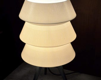 May include: A modern table lamp with three stacked, pleated lampshades. The shades are a light cream color and sit atop a dark gray tripod base. The lamp is illuminated, casting a warm glow.