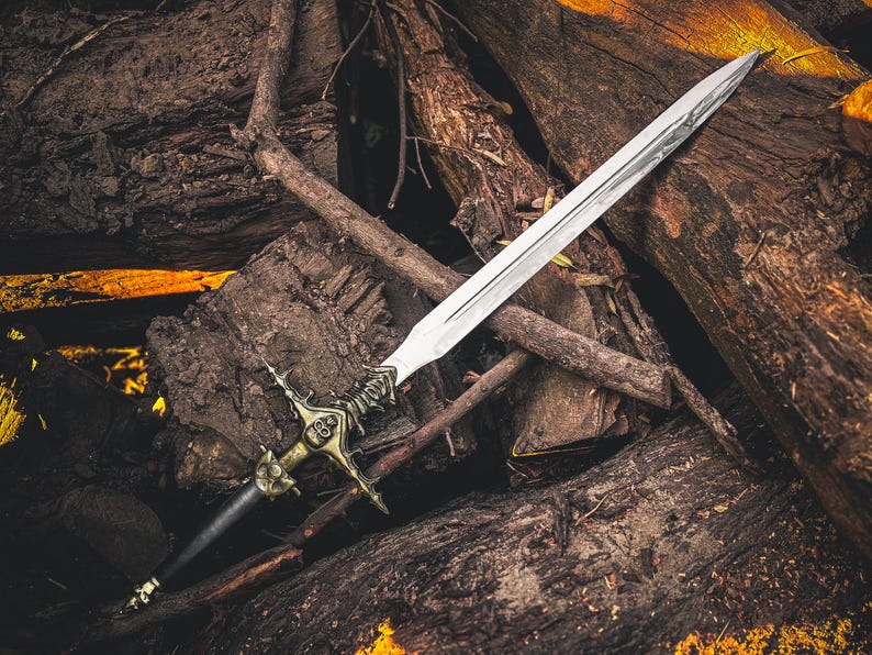 May include: A silver sword with an ornate gold and black hilt rests on a pile of weathered logs. The blade is long and sharp, reflecting light. The hilt features intricate details, suggesting a fantasy or historical theme.