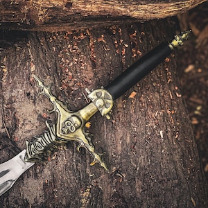 May include: A decorative sword with a silver blade and a detailed gold-colored hilt featuring skull and ornate designs. The black handle contrasts with the metallic elements. The sword rests on a textured, brown wooden surface.