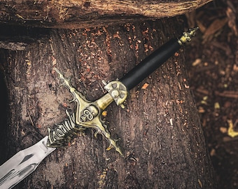 May include: A decorative sword with a silver blade and a detailed gold-colored hilt featuring skull and ornate designs. The black handle contrasts with the metallic elements. The sword rests on a textured, brown wooden surface.