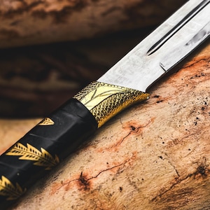 K&ouml;nnte beinhalten: Nahaufnahme eines dekorativen Messers. Die silberne Klinge kontrastiert mit dem schwarzen Griff, der mit goldenen Blattmustern verziert ist. Ein goldfarbener Knauf mit Schuppenmuster verbindet Klinge und Griff. Das Messer liegt auf einer Holzoberfl&auml;che.