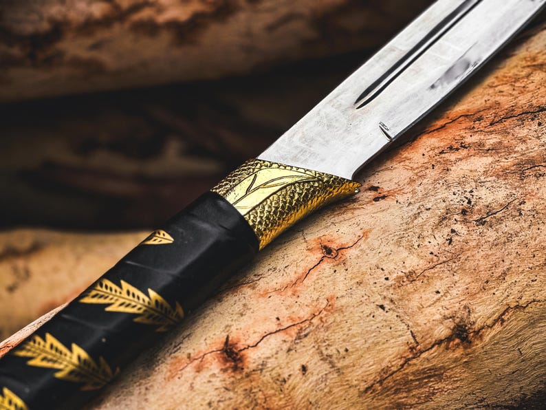 K&ouml;nnte beinhalten: Nahaufnahme eines dekorativen Messers. Die silberne Klinge kontrastiert mit dem schwarzen Griff, der mit goldenen Blattmustern verziert ist. Ein goldfarbener Knauf mit Schuppenmuster verbindet Klinge und Griff. Das Messer liegt auf einer Holzoberfl&auml;che.