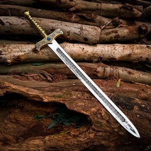 May include: An ornate sword with a gold-colored hilt and a silver blade, resting on a bed of wooden logs. The hilt features intricate designs and a blue gem. The blade has a detailed pattern. The sword is a fantasy or historical item.