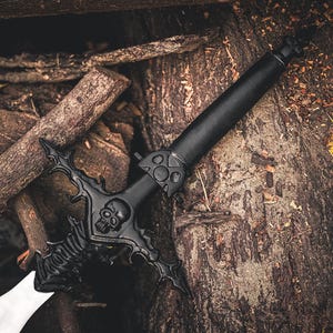 May include: A black fantasy sword with a skull design on the hilt and a white blade. The sword is set against a backdrop of wood logs and yellow foliage, creating a dramatic contrast.