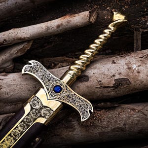 May include: A decorative sword with a gold-colored handle and blade, featuring intricate engraved patterns. A blue gem is set in the center of the hilt. The sword rests on a bed of wooden logs, creating a rustic aesthetic.