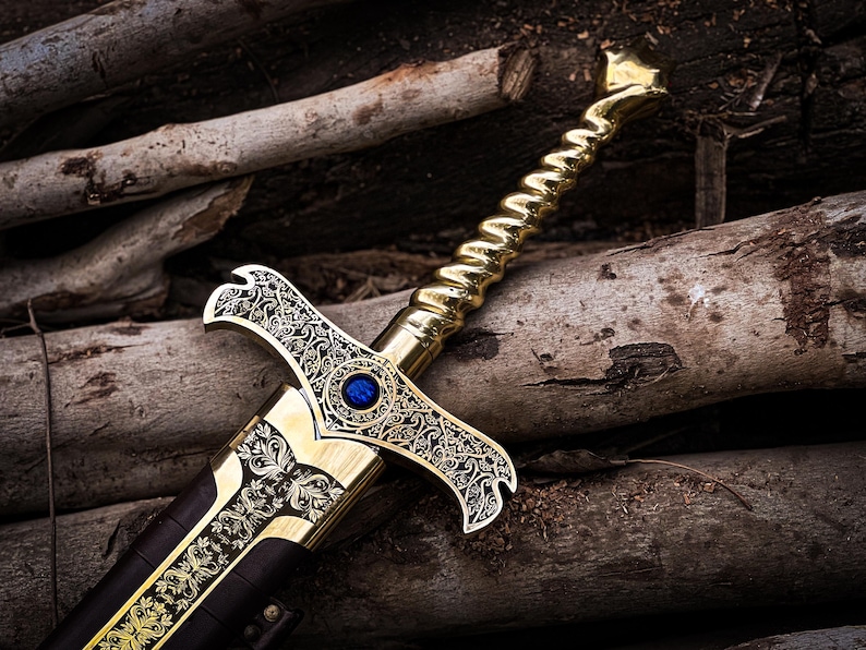 May include: A decorative sword with a gold-colored handle and blade, featuring intricate engraved patterns. A blue gem is set in the center of the hilt. The sword rests on a bed of wooden logs, creating a rustic aesthetic.