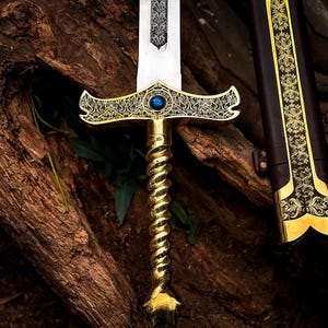May include: A decorative sword with a gold-colored handle and crossguard, featuring intricate patterns and a blue gem. The blade is silver, and the scabbard is brown with gold accents. The sword is displayed against a textured, brown background.