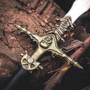 May include: A detailed image of a decorative sword with a bronze-colored hilt and a silver blade. The hilt features intricate skull and ornate designs. The sword rests on a bed of dark brown wood and branches.