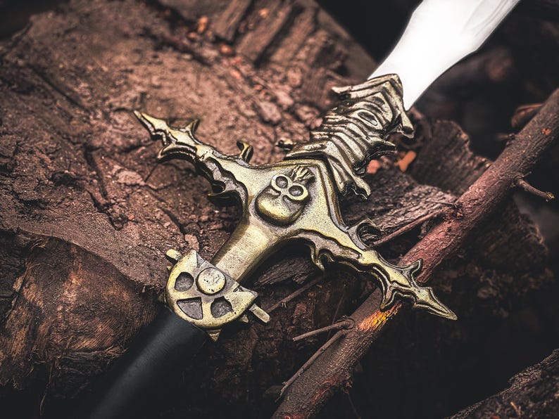 May include: A detailed image of a decorative sword with a bronze-colored hilt and a silver blade. The hilt features intricate skull and ornate designs. The sword rests on a bed of dark brown wood and branches.