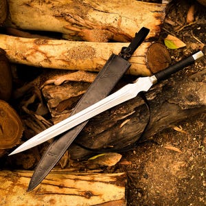 May include: A silver sword with a black handle and a black leather sheath rests on a pile of logs. The sword has a double-edged blade and a decorative hilt. The sheath is open, revealing the blade.