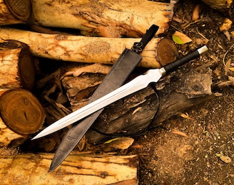 May include: A silver sword with a black handle and a black leather sheath rests on a pile of logs. The sword has a double-edged blade and a decorative hilt. The sheath is open, revealing the blade.