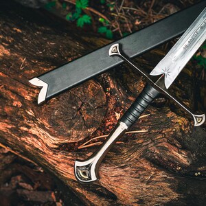 May include: A detailed image of a sword with a silver blade and a black handle, resting on a weathered wooden surface. The sword has a decorative hilt and a black sheath. The blade has an inscription and a star design.