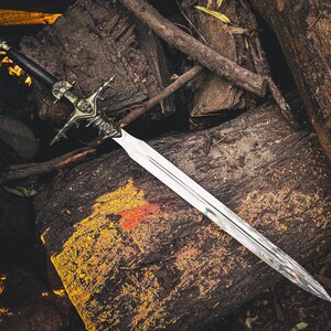 May include: A detailed image of a decorative sword with a silver blade and ornate hilt. The hilt features intricate designs and a dark handle. The sword rests on a bed of weathered wood, creating a rustic aesthetic.