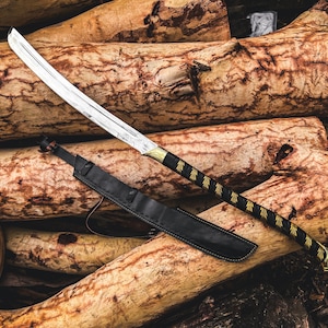 K&ouml;nnte beinhalten: Ein Katana-Schwert mit silberner Klinge und schwarzem Griff mit goldenen Akzenten liegt auf einem Stapel Holzscheite. Eine schwarze Lederscheide liegt neben dem Schwert. Der Griff des Schwertes hat ein dekoratives Muster.