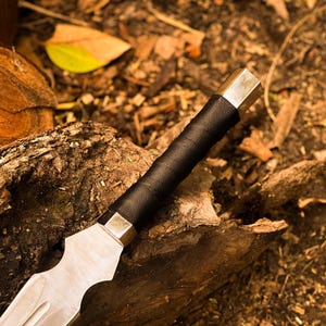 Peut inclure: Gros plan sur un poignard argent&eacute; et noir. La lame a un design incurv&eacute; unique. Le manche est envelopp&eacute; de cuir noir et a une extr&eacute;mit&eacute; en m&eacute;tal argent&eacute;. Le poignard repose sur un morceau de bois.