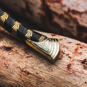 K&ouml;nnte beinhalten: Nahaufnahme eines dekorativen Schwertgriffs. Der Griff ist goldfarben mit einem verzierten Design, und der Griff ist schwarz mit goldenen Blattmustern. Das Schwert liegt auf einem St&uuml;ck verwittertem Holz.