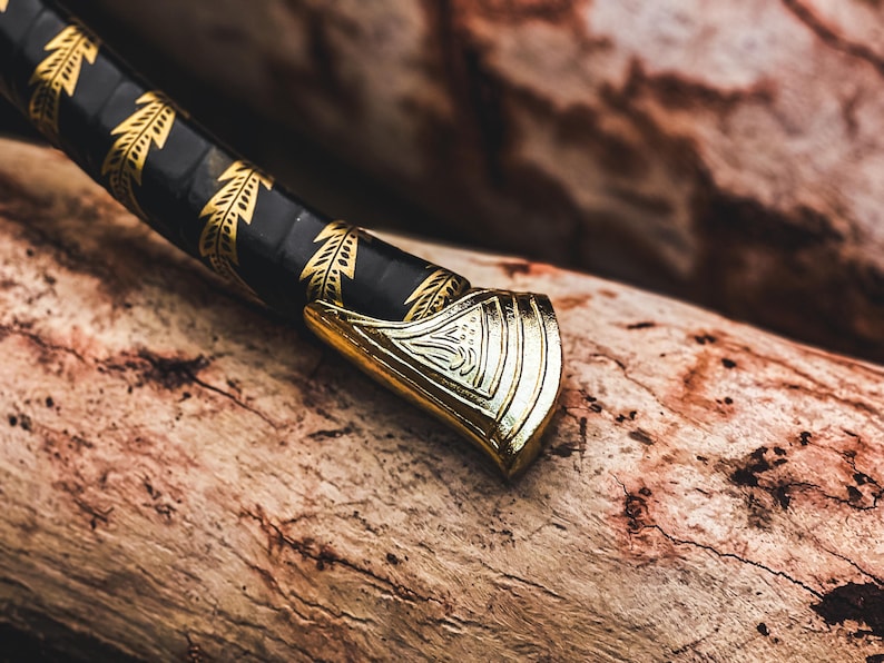 K&ouml;nnte beinhalten: Nahaufnahme eines dekorativen Schwertgriffs. Der Griff ist goldfarben mit einem verzierten Design, und der Griff ist schwarz mit goldenen Blattmustern. Das Schwert liegt auf einem St&uuml;ck verwittertem Holz.