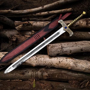 May include: A silver and gold sword with a decorative hilt and a blue gem, resting on a pile of logs. The sword has a detailed blade and is accompanied by a red leather sheath.