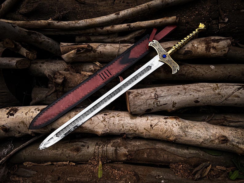 May include: A silver and gold sword with a decorative hilt and a blue gem, resting on a pile of logs. The sword has a detailed blade and is accompanied by a red leather sheath.