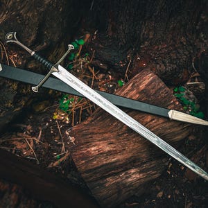 May include: A silver sword with a black handle and a black sheath rests on a wooden surface. The sword has a crossguard and a pointed tip. The blade has etched text. The sheath has a silver tip.