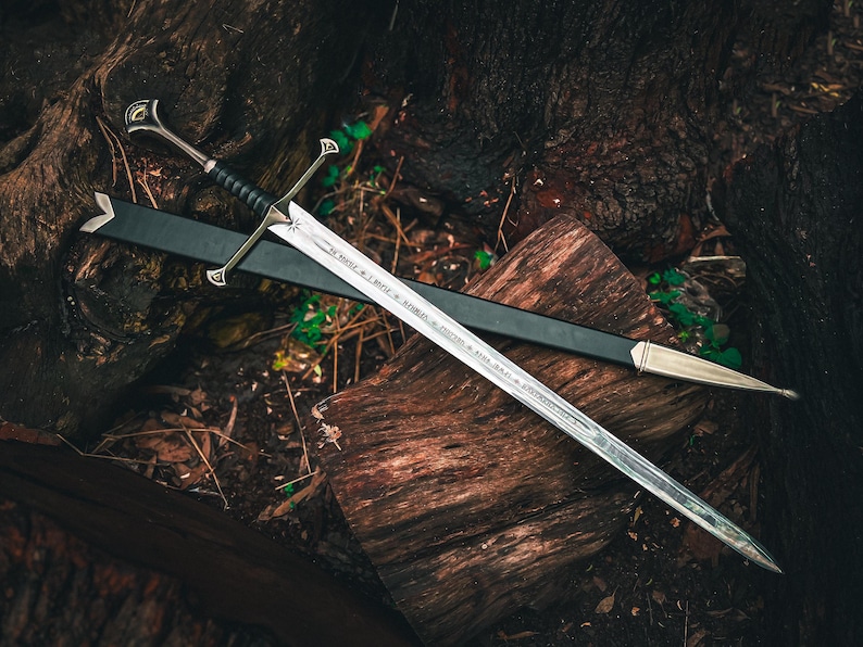 May include: A silver sword with a black handle and a black sheath rests on a wooden surface. The sword has a crossguard and a pointed tip. The blade has etched text. The sheath has a silver tip.
