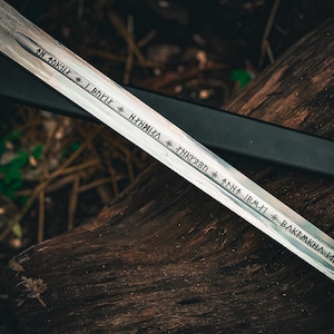 May include: A silver sword with etched inscriptions and a starburst design on the hilt. The blade is resting on a weathered wooden surface, with a black sheath visible in the background. The sword's inscriptions are in a stylized font.