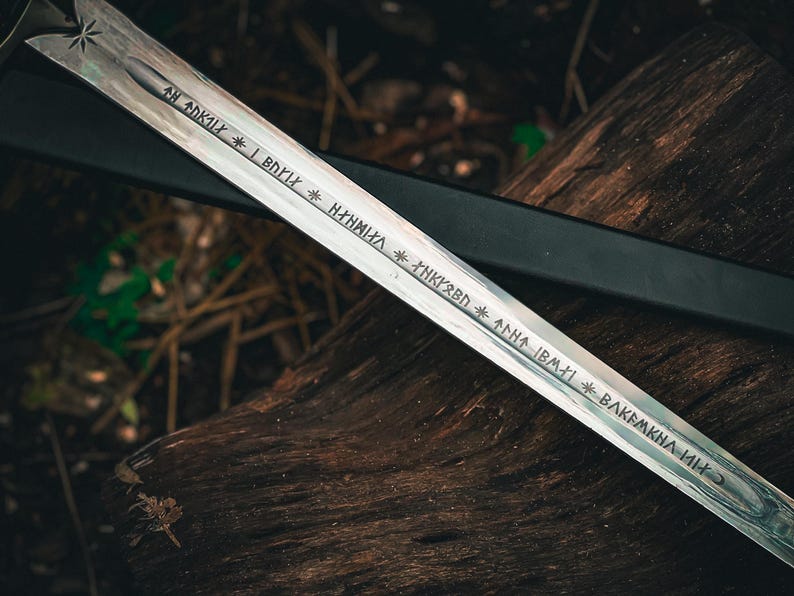 May include: A silver sword with etched inscriptions and a starburst design on the hilt. The blade is resting on a weathered wooden surface, with a black sheath visible in the background. The sword's inscriptions are in a stylized font.