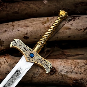 May include: A decorative sword with a gold-colored handle and crossguard, featuring intricate patterns and a blue gem. The blade is silver with a detailed design. The sword rests on a background of wooden logs.