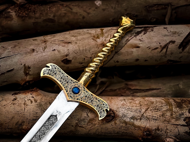 May include: A decorative sword with a gold-colored handle and crossguard, featuring intricate patterns and a blue gem. The blade is silver with a detailed design. The sword rests on a background of wooden logs.
