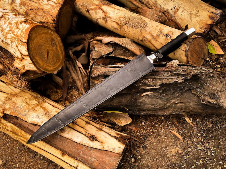 Peut inclure: Un long couteau noir avec un &eacute;tui en cuir repose sur une pile de b&ucirc;ches. Le couteau a une mitre argent&eacute;e et un manche noir. L'&eacute;tui est gris fonc&eacute; avec des coutures d&eacute;coratives. La sc&egrave;ne se d&eacute;roule en ext&eacute;rieur avec une lumi&egrave;re naturelle.