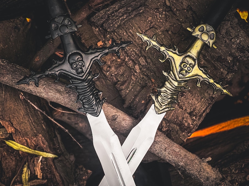 May include: Two decorative swords with skull-shaped hilts and ornate designs. One sword is black, the other is gold, both with silver blades. The swords are crossed against a backdrop of wooden branches.