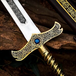 May include: A detailed close-up of a decorative sword with a silver blade and ornate gold hilt. The hilt features a blue gem and intricate patterns. A brown leather sheath with gold accents is visible in the background.