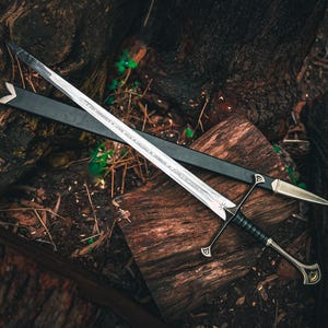 May include: A silver sword with a black handle and crossguard, resting on a wooden surface. The sword has a long, double-edged blade and is accompanied by a black sheath. The handle is wrapped in black material, and the crossguard has decorative elements.