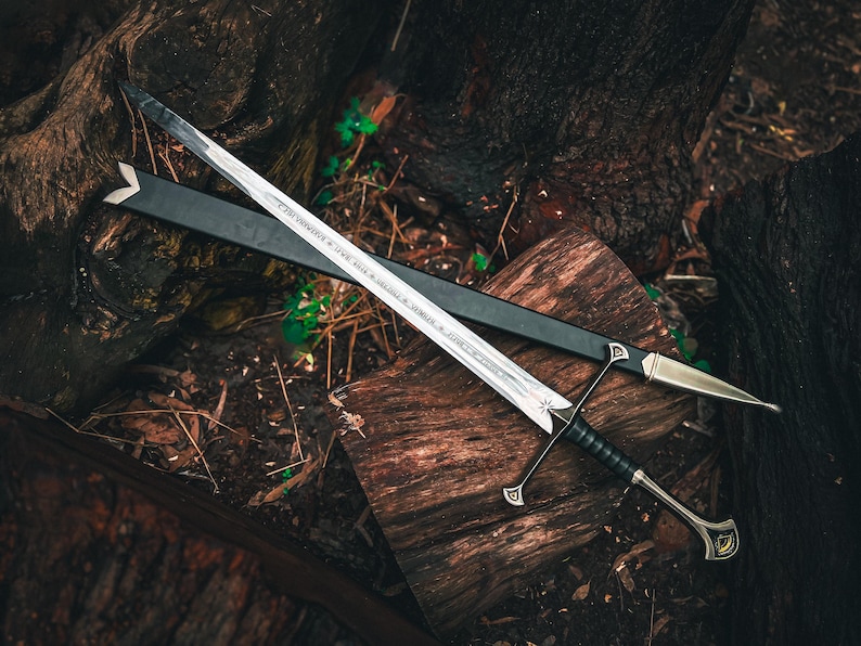 May include: A silver sword with a black handle and crossguard, resting on a wooden surface. The sword has a long, double-edged blade and is accompanied by a black sheath. The handle is wrapped in black material, and the crossguard has decorative elements.