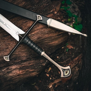 May include: A detailed image of a sword and scabbard resting on a wooden surface. The sword features a silver blade with etched text and a star design. The handle is wrapped in black leather, and the scabbard is black with silver accents.